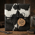 Cow print wrapping paper with a leather cord bow and "For the Wild One" tag on a wrapped present.