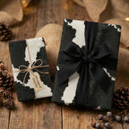 Rustic gifts wrapped in black and white cow print wrapping paper with twine and black ribbon on wood table.